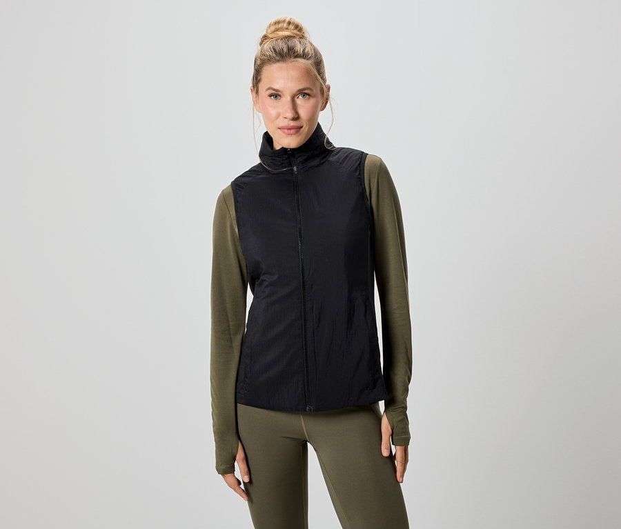 Egy szőke hajú nő fekete mellényt visel egy zöld hosszú ujjú felső és leggings felett.