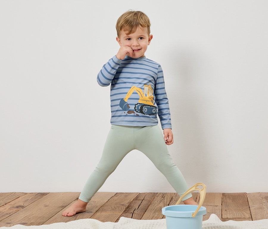 Egy fiú mezítláb áll egy fapadlón, egy markolós, csíkos, hosszú ujjú pólót és világoszöld leggingset visel. Előtte egy kék vödör áll.