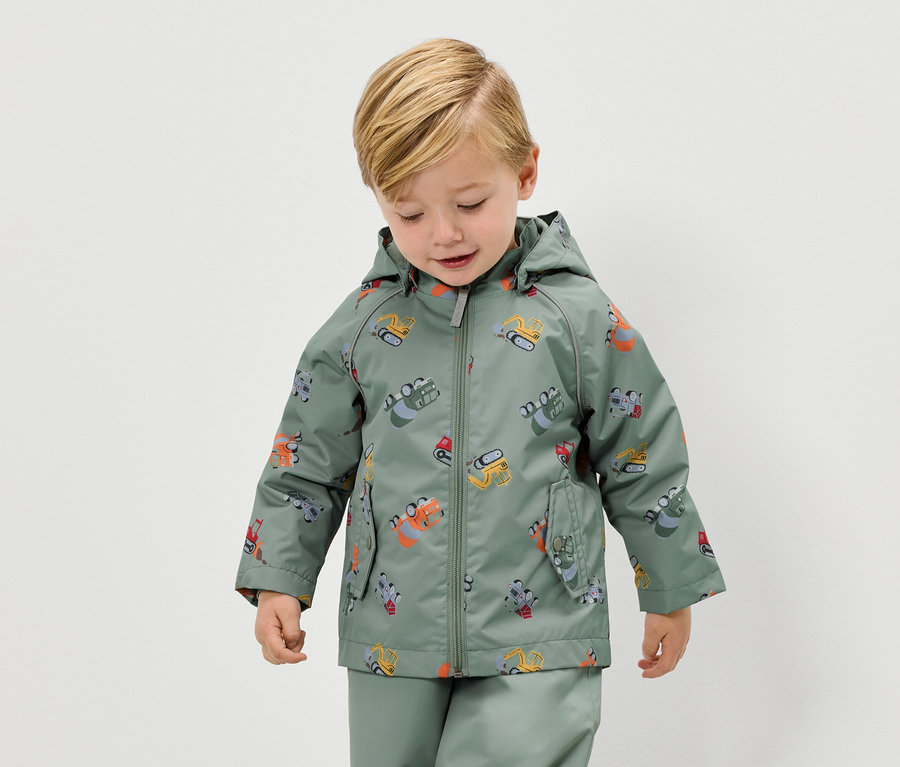 Egy szőke fiú zöld, markoló mintás esőkabátot és hozzáillő esőnadrágot visel.