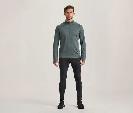 Férfi áll sötétszürke, hosszú ujjú, cipzáras sportfelsőben, hüvelykujjnyílásokkal, fekete sportleggingsben és fekete sportcipőben.