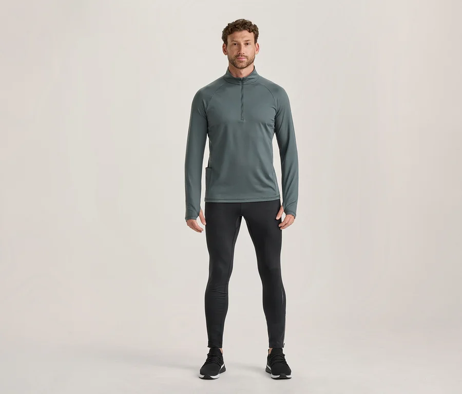Férfi áll sötétszürke, hosszú ujjú, cipzáras sportfelsőben, hüvelykujjnyílásokkal, fekete sportleggingsben és fekete sportcipőben.