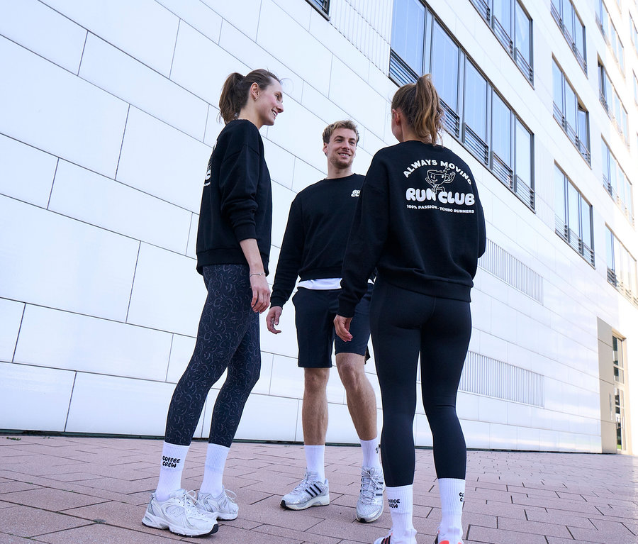 Három ember, két nő és egy férfi, állva mosolyogva beszélget. Mindannyian fekete szabadidőfelsőt és fehér Uniszex Coffe Crew teniszzoknit viselnek. A férfi fekete sport rövidnadrágot visel. Az egyik nő antracit színű, leopárdmintás női sportnadrágot, a másik fekete női sportnadrágot visel.