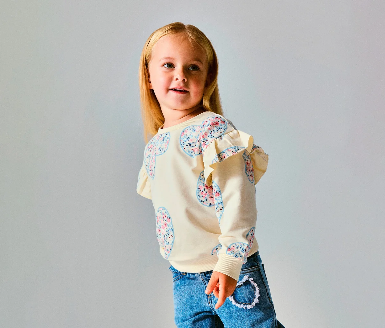 Fiatal szőke hajú lány krém színű NAME IT Kinder-Sweater pulóvert visel virágos szívmintákkal és fodrokkal a vállán, kék farmerrel, jobbra néz.