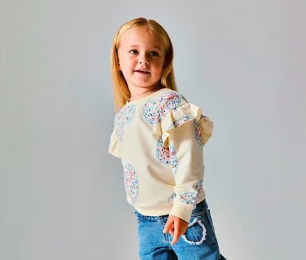 Fiatal szőke hajú lány krém színű NAME IT Kinder-Sweater pulóvert visel virágos szívmintákkal és fodrokkal a vállán, kék farmerrel, jobbra néz.