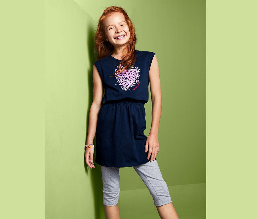 Mosolygó lány pózol sötétkék, flitteres lány ruhában és szürke leggingsben.