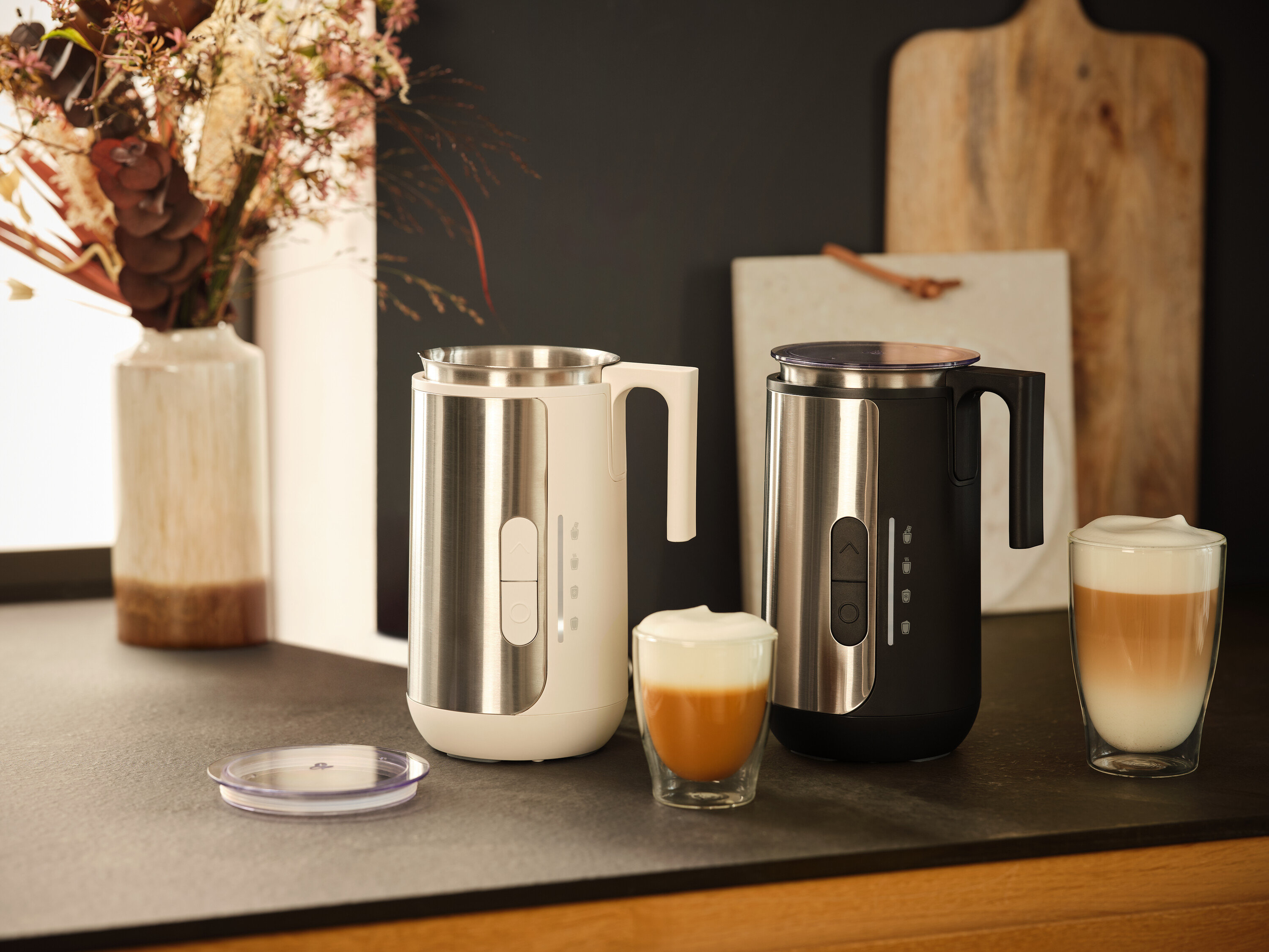 Két tejhabosító, egy fehér és egy fekete, áll egy pulton két pohár tejhabbal.