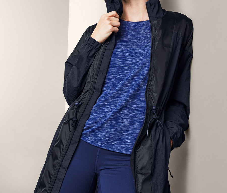 A falnak támaszkodó szőke nő funkcionális kabátot, kék pólót és leggingset visel.