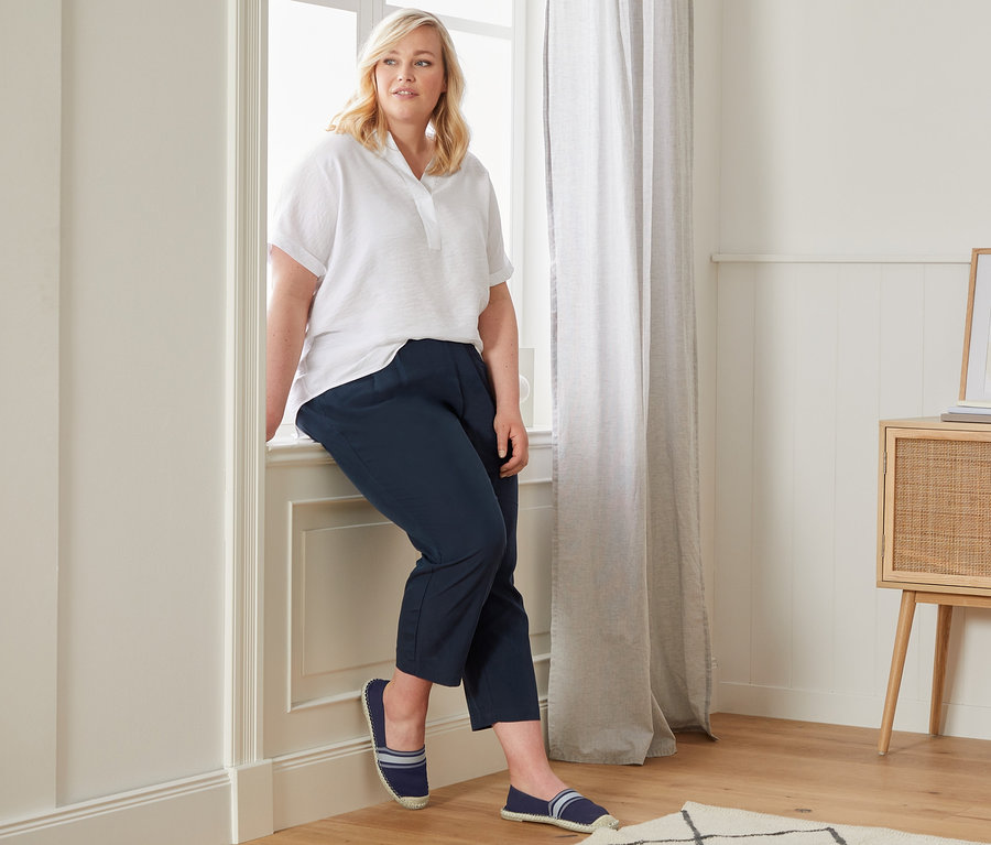 Nő ül az ablakpárkányon, fehér oversize blúzt, sötétkék 7/8-os chino nadrágot és sötétkék espadrilles-t visel.
