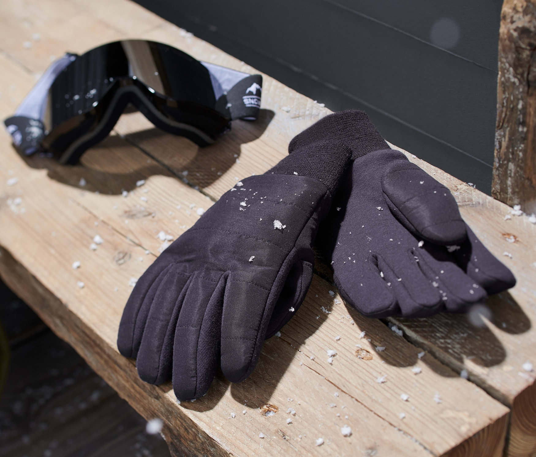 Fekete kötött kesztyű és sí szemüveg egy fa padon.