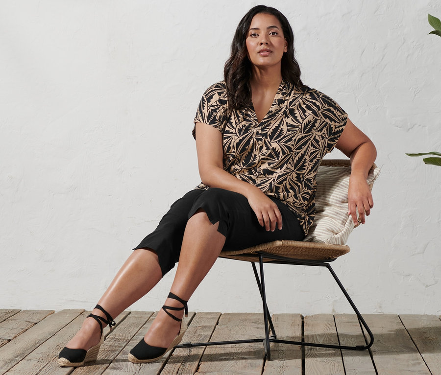 Nő ül egy széken, és ékettsarkú espadrilles-t, 3/4-es sztreccsnadrágot és nyomott mintás, állógalléros felsőt visel.