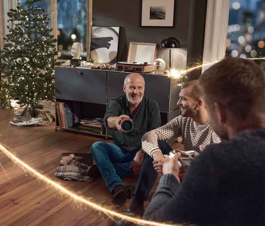 Három férfi ül a nappaliban a földön, egyikük egy Bluetooth hangszórót tart műbőr borítással. Férfi flaneling, szürke, Férfi farmernadrág, kék és Férfi kötött pulóver, mintás van rajtuk.