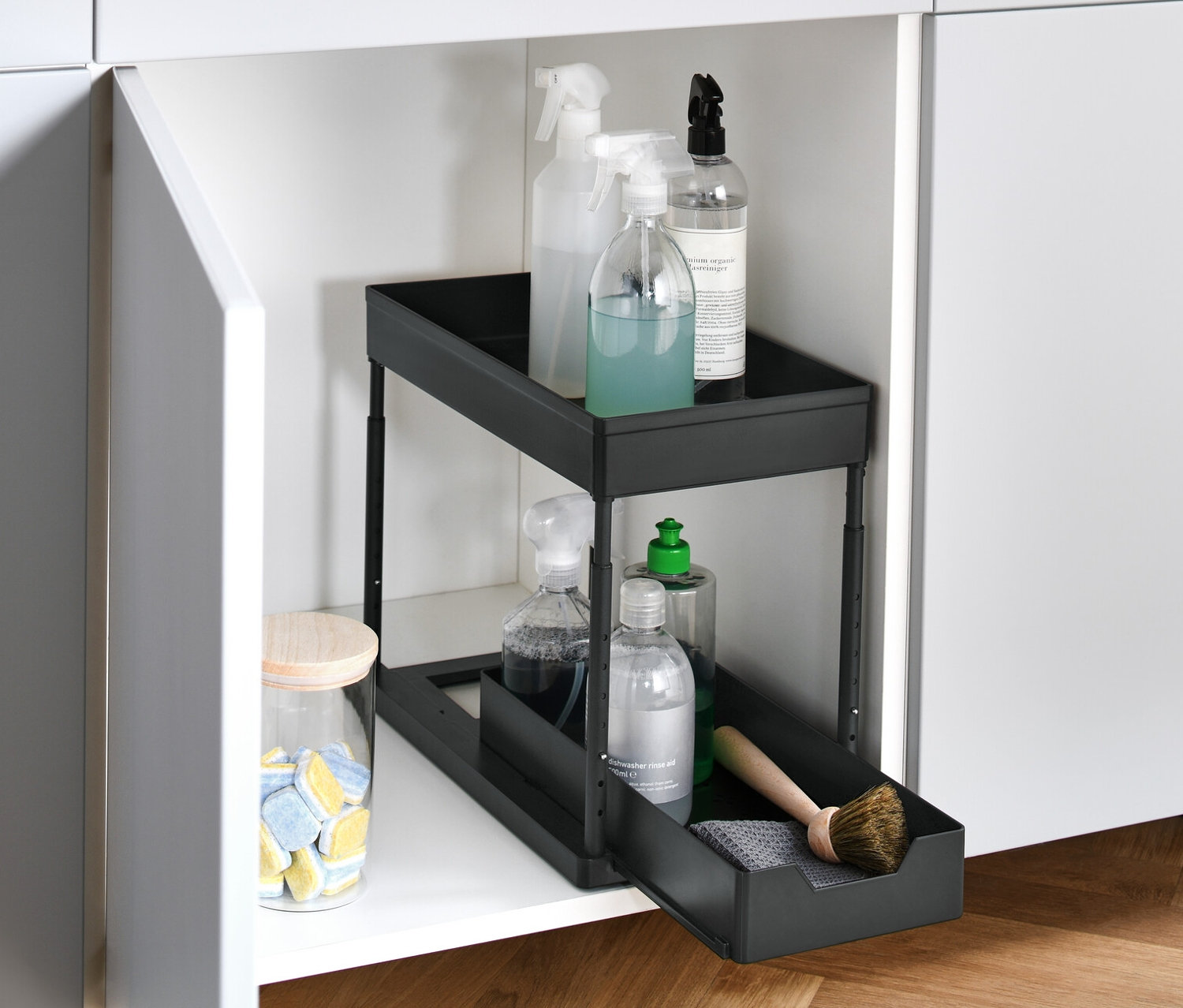 Egy nyitott, fehér szekrény állítható magasságú kihúzható tárolóval, benne tisztítószerekkel.