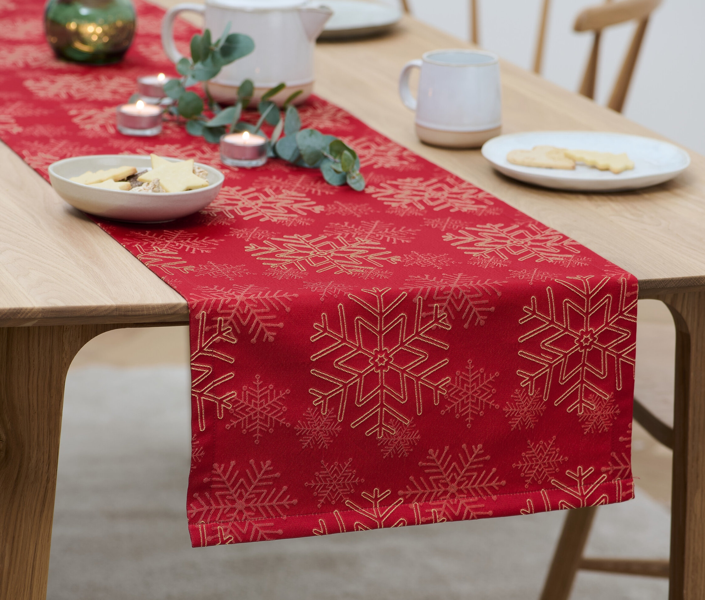 Egy fa asztalon piros, hópehely mintás jacquard asztali futó található, rajta süteményes tányérok, gyertyák és egy teáskanna.