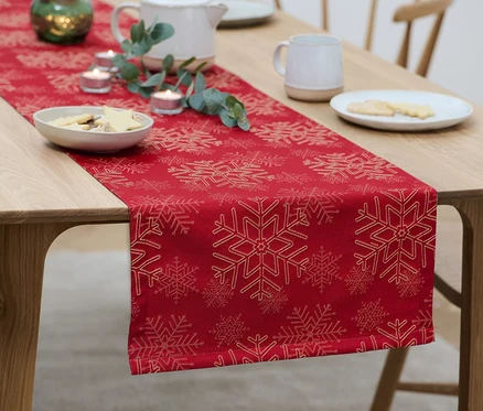 Egy fa asztalon piros, hópehely mintás jacquard asztali futó található, rajta süteményes tányérok, gyertyák és egy teáskanna.