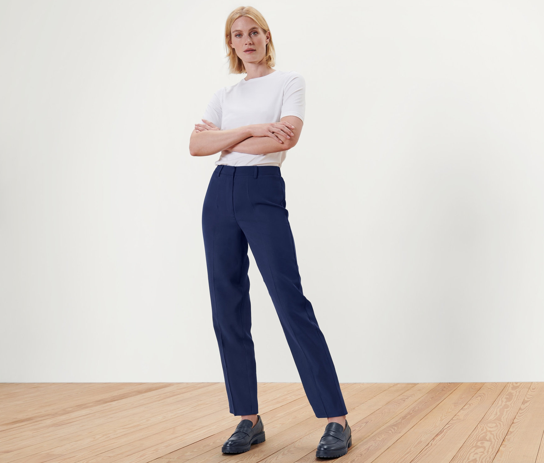 Szőke nő áll keresztbe tett karokkal, kék szövött nadrágban és cipőben a fapadlón.