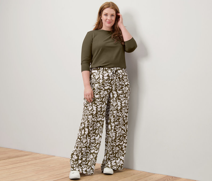 Egy nő pózol egy khaki 3/4-es ujjú felsőben és egy nyomott mintás belebújós nadrágban.
