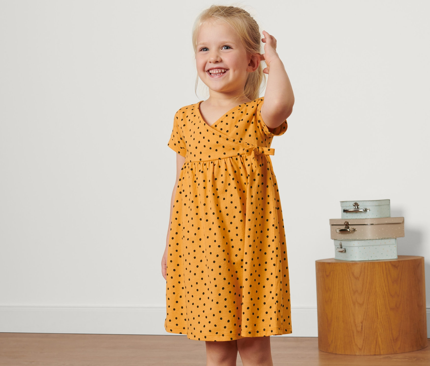 Egy mosolygós szőke kislány sárga, fekete pöttyös gyerek jersey ruhát visel.