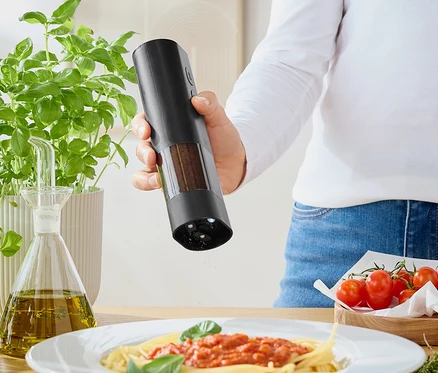 Nő fűszereket szór egy darálóból a paradicsomszószos spagettire. Az asztalon bazsalikom, olívaolaj és paradicsom található.