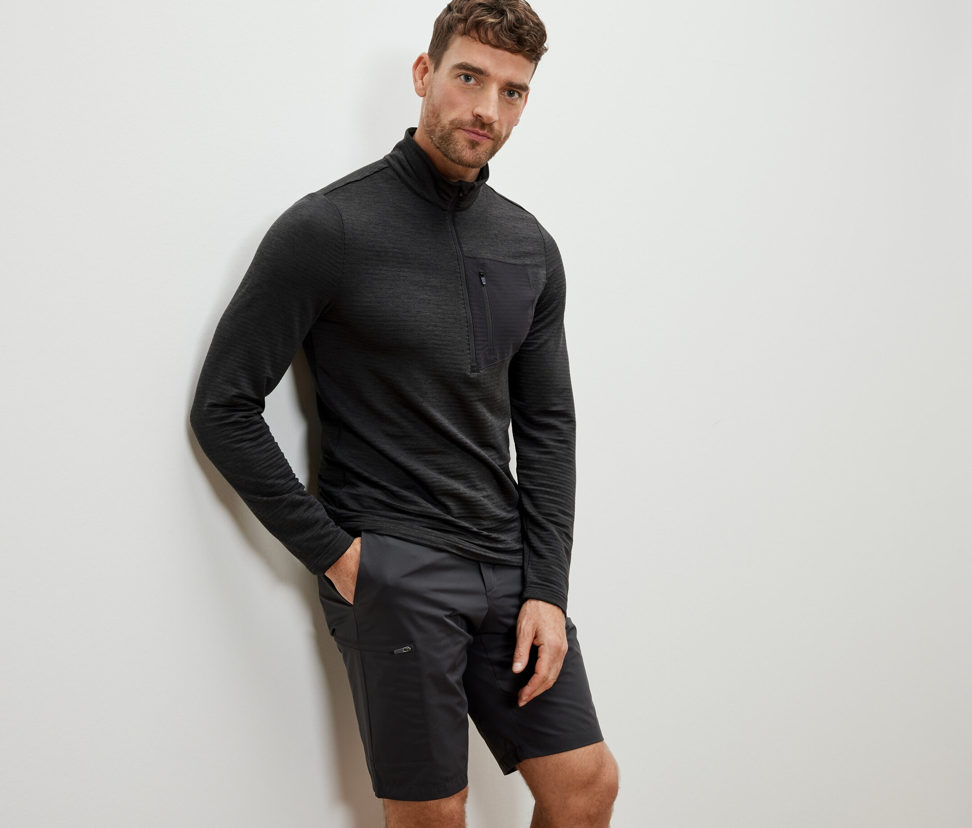 Férfi a falnak dőlve, antracit színű hosszú ujjú funkcionális felsőt és rövidnadrágot visel.