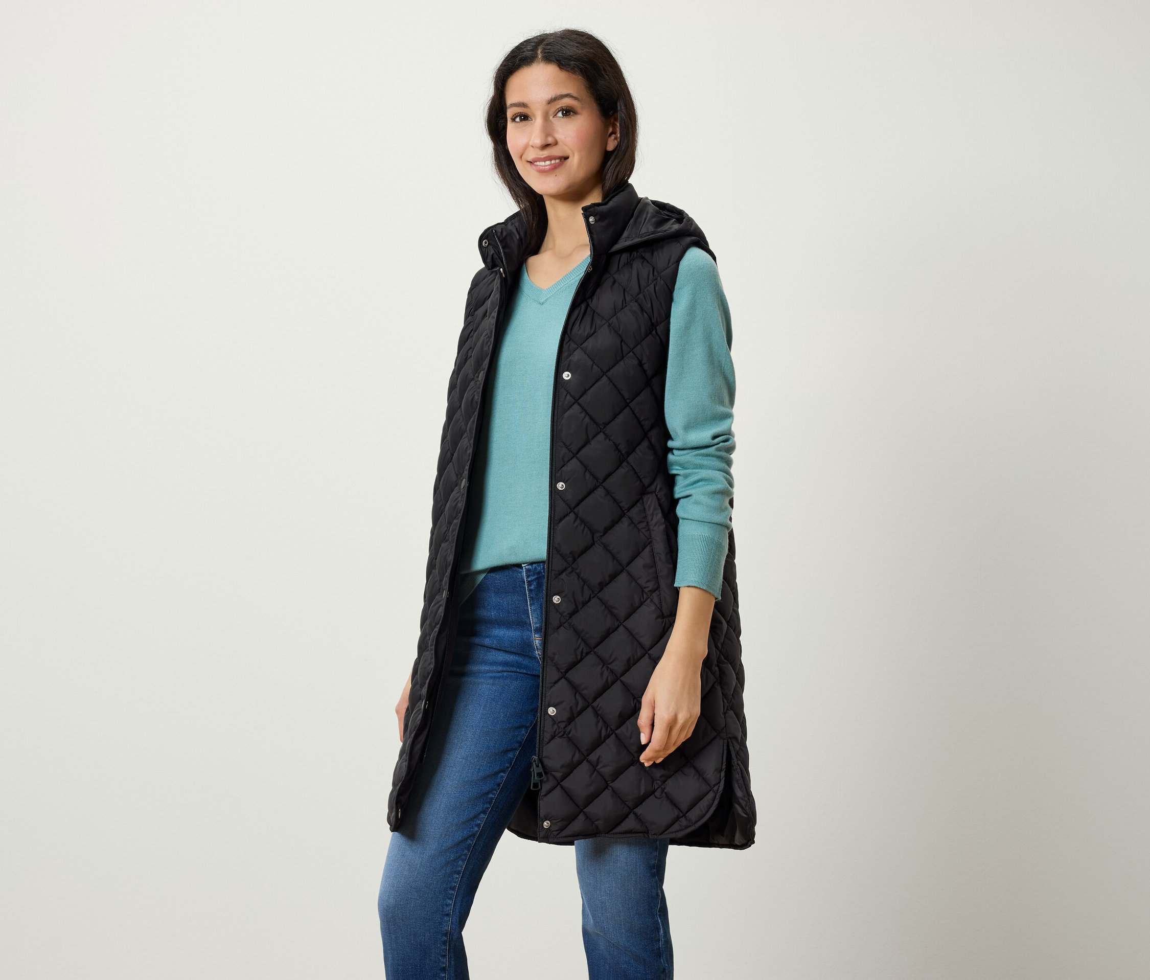 Egy nő hosszú, fekete, steppelt, kapucnis mellényt, kék pulóvert és farmert visel.