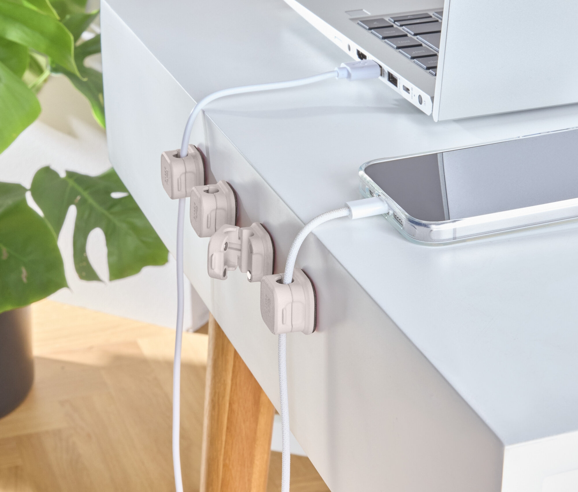 Ezüst laptop és telefon töltődik egy fehér asztalon, fa kábelrögzítőkkel.