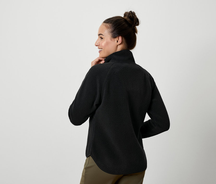 Egy nő a válla fölött néz hátra, fekete fleece felsőt és khaki nadrágot visel.