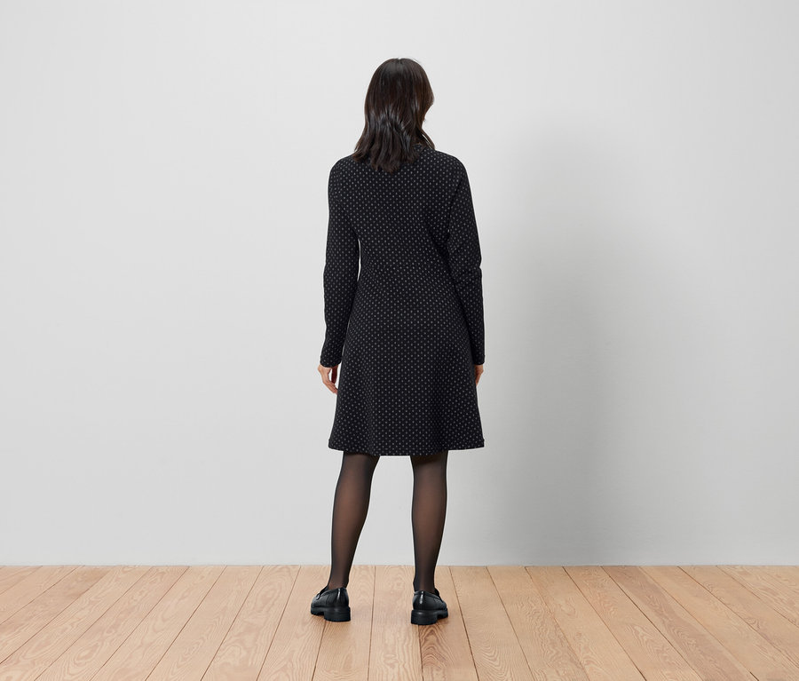 Egy nő háttal áll a kamerának, fekete ruhát és harisnyát visel.