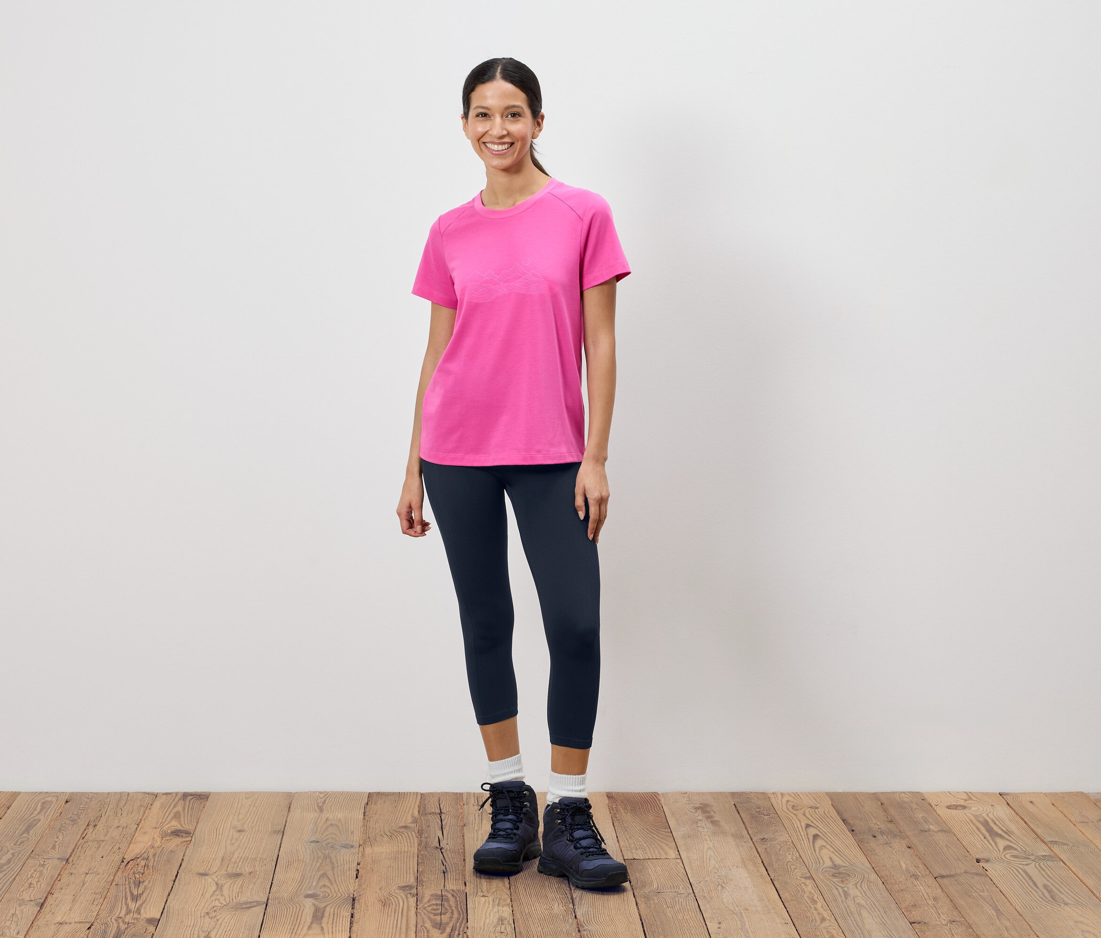 Nő áll egy fehér fal előtt, rózsaszín pólót, sötét leggingset és túrabakancsot visel.