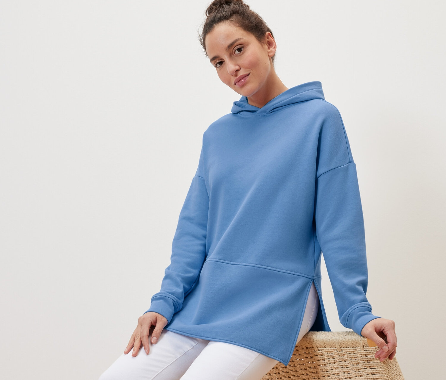 Nő ül egy fonott széken, világoskék, hosszú kapucnis felsőt visel.