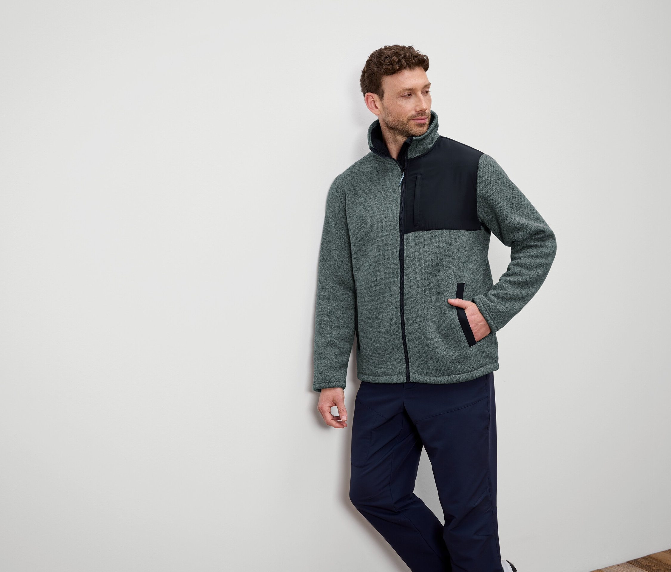 Egy falnak támaszkodó férfi szürke polárkabátot visel fekete vállakkal és sötétkék nadrágot.