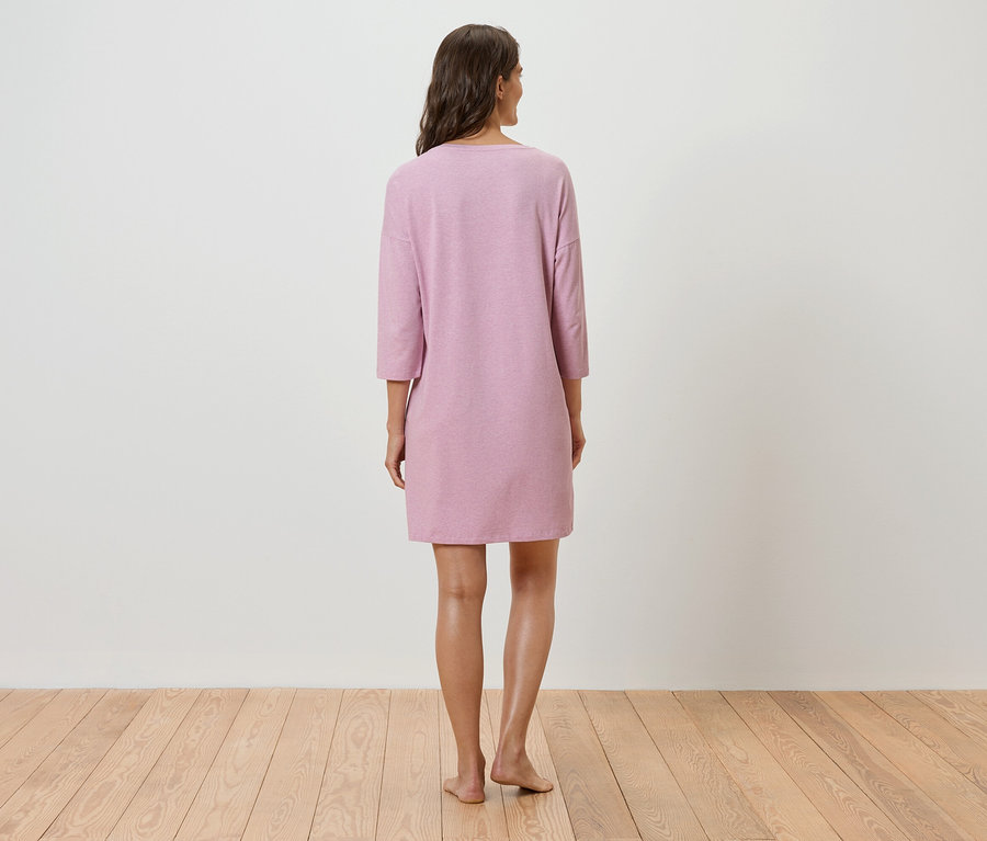 Egy nő áll háttal a kamerának, rózsaszín, háromnegyedes ujjú ruhát visel.