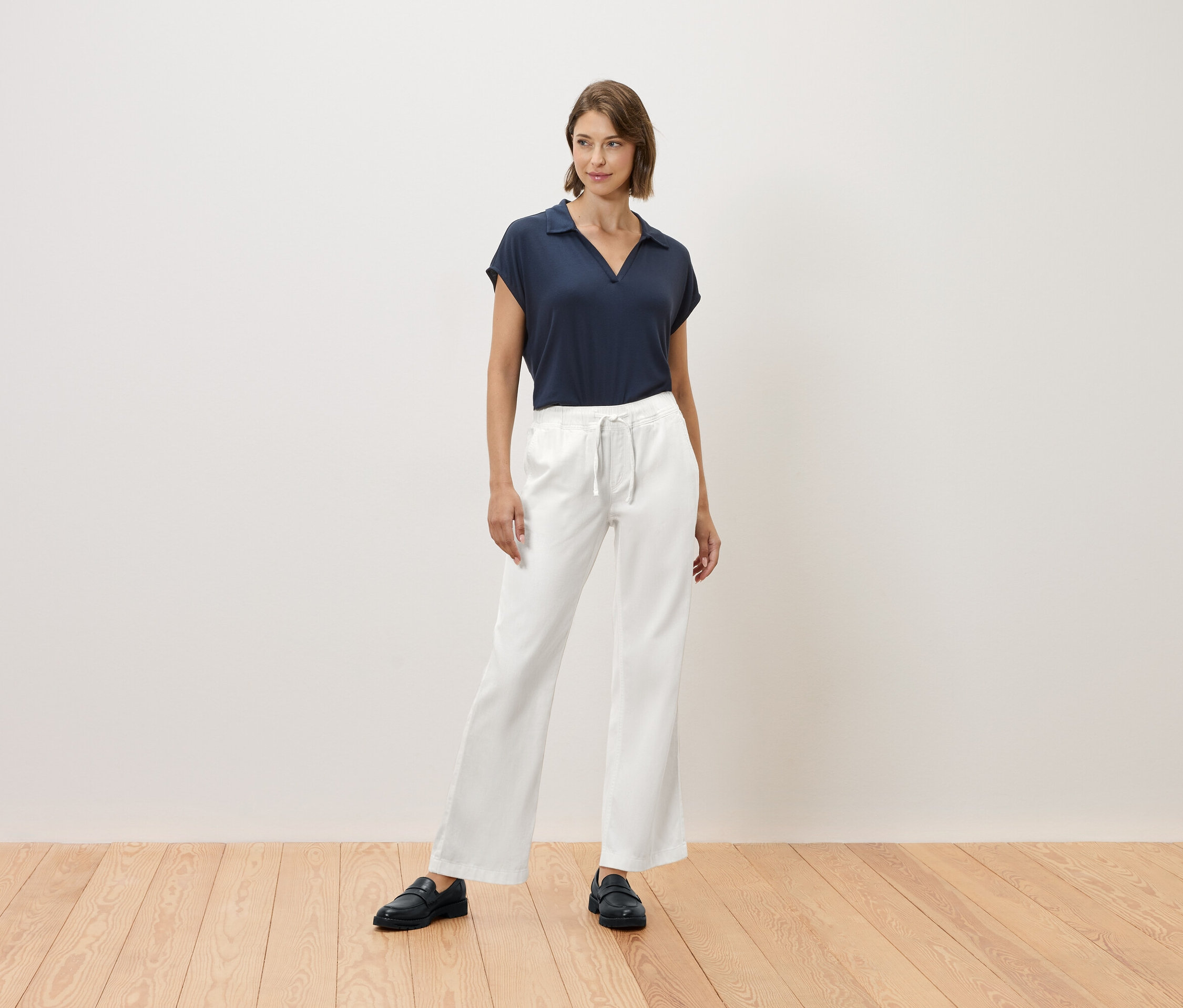 Egy nő áll egy fapadlón, sötétkék pólót és fehér nadrágot visel.