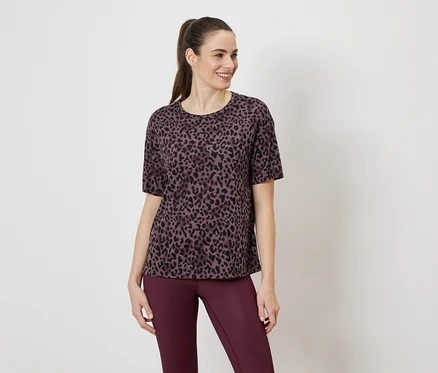 Nő lila leopárdmintás pólót és lila leggingset visel fehér háttér előtt.