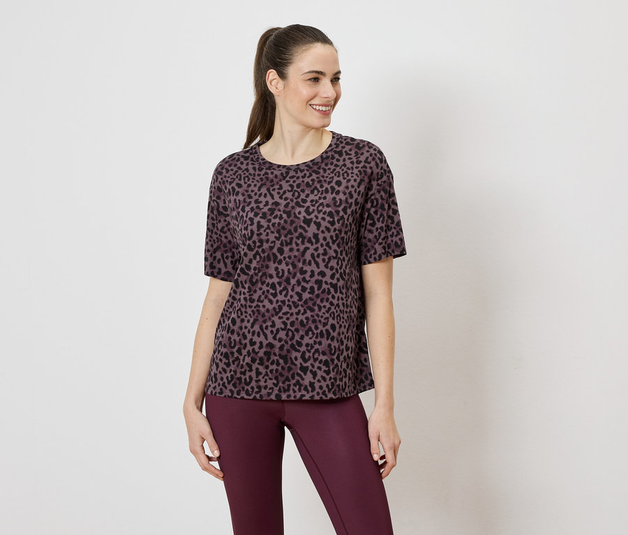 Nő lila leopárdmintás pólót és lila leggingset visel fehér háttér előtt.
