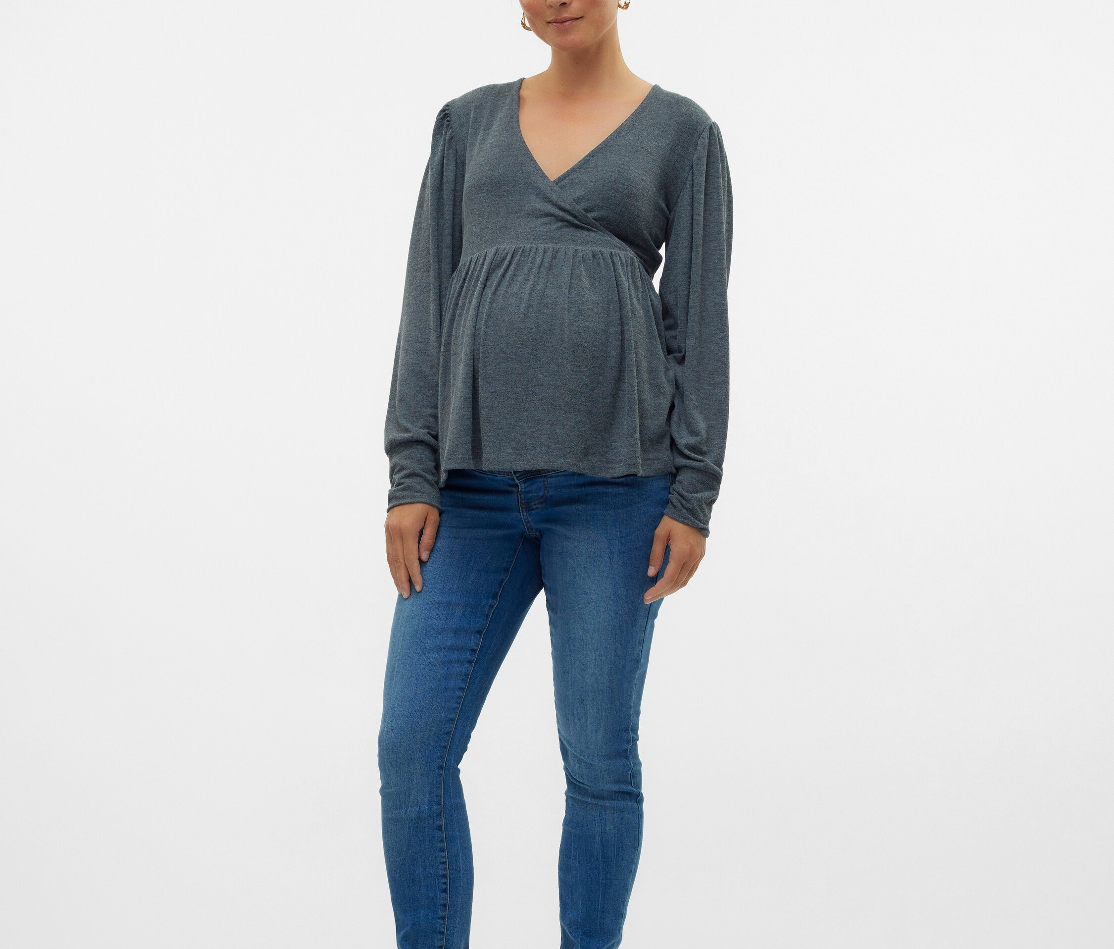 Egy várandós nő szürke, hosszú ujjú felsőt, kék farmert és fekete balerinacipőt visel.