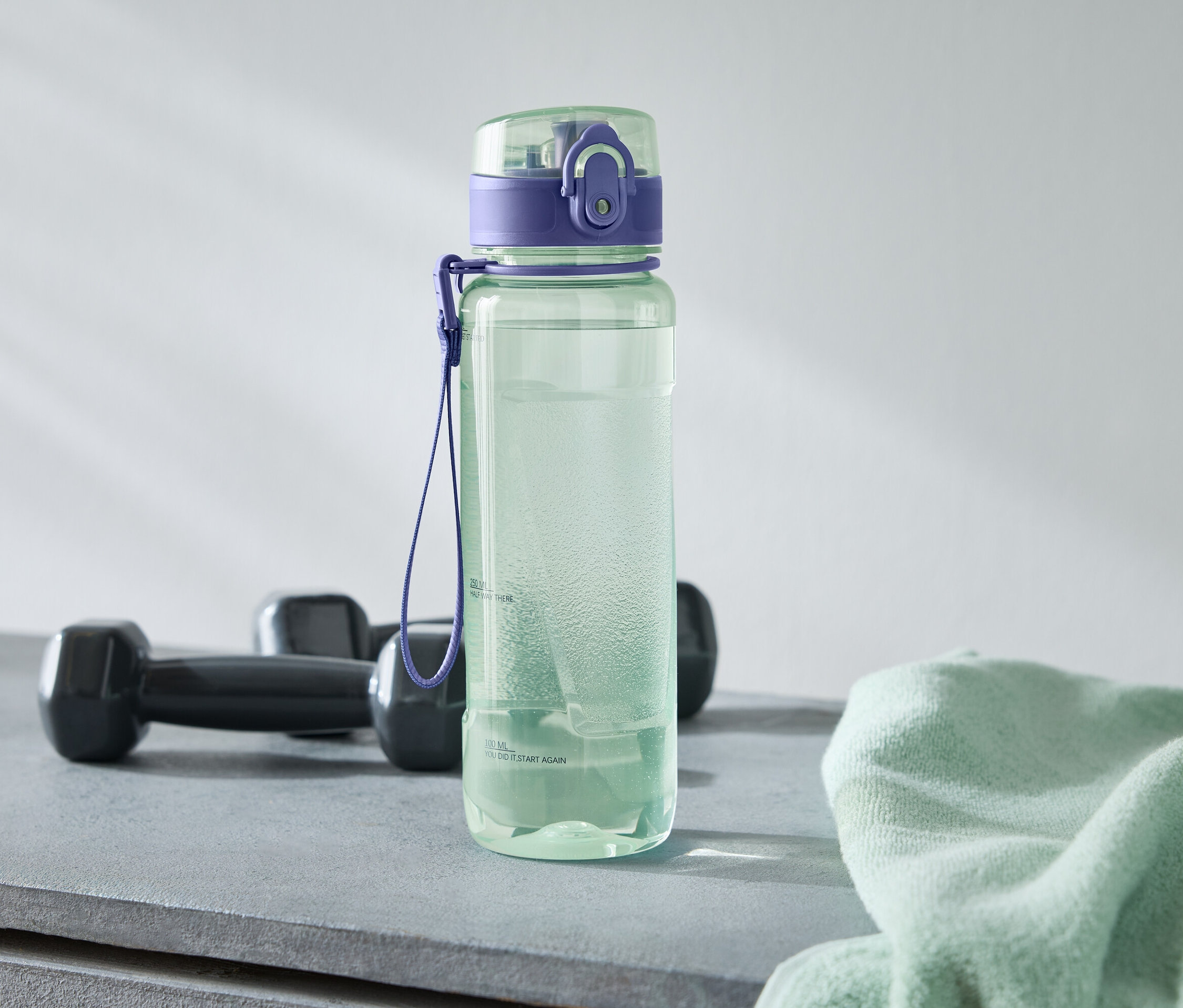 Egy zöld kulacs lila kupakkal áll két fekete súlyzó és egy világoszöld törölköző mellett egy szürke felületen.
