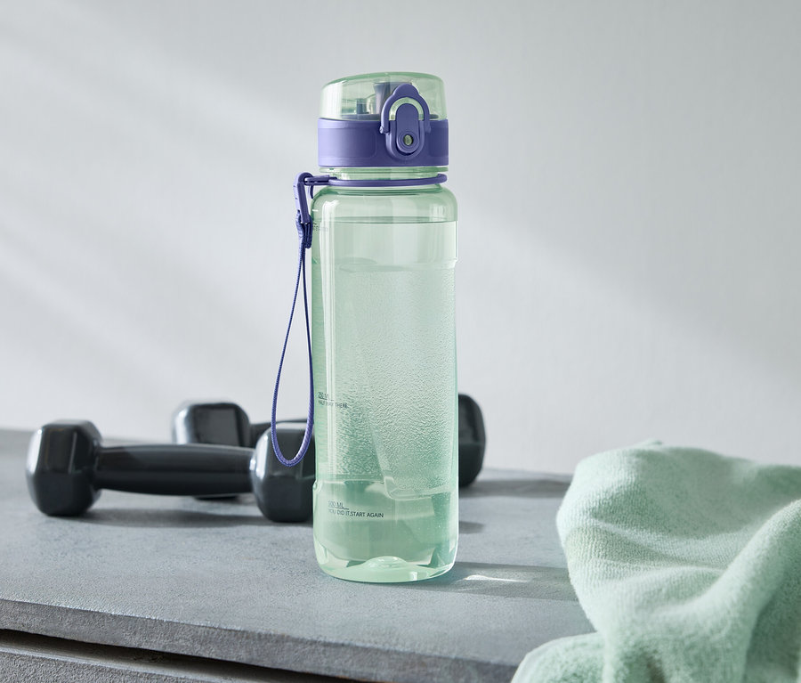 Egy zöld kulacs lila kupakkal áll két fekete súlyzó és egy világoszöld törölköző mellett egy szürke felületen.