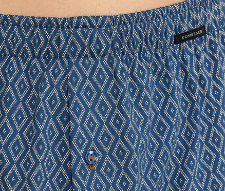 Közeli felvétel egy kék SCHIESSER jersey boxeralsóról, fehér rombuszmintával, rugalmas derékpánttal, fekete SCHIESSER címkével és narancssárga varrású kék gombbal.