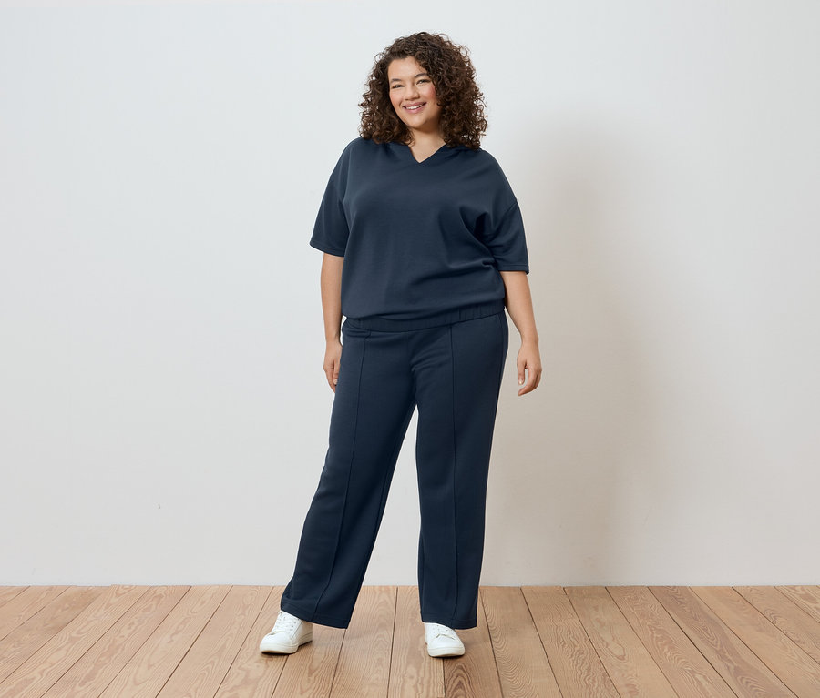 Göndör hajú nő sötétkék pólóban és nadrágban, fehér fal előtt.