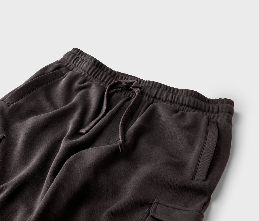 Egy sötétbarna színű, húzózsinóros, gumis derekú és zsebes jogging alsó közeli képe szürke háttér előtt.