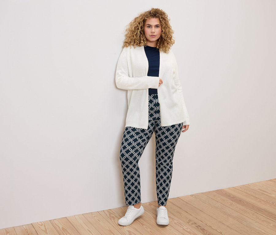 Göndör hajú nő fehér kardigánban, sötétkék felsőben és mintás nadrágban, fehér fal előtt.