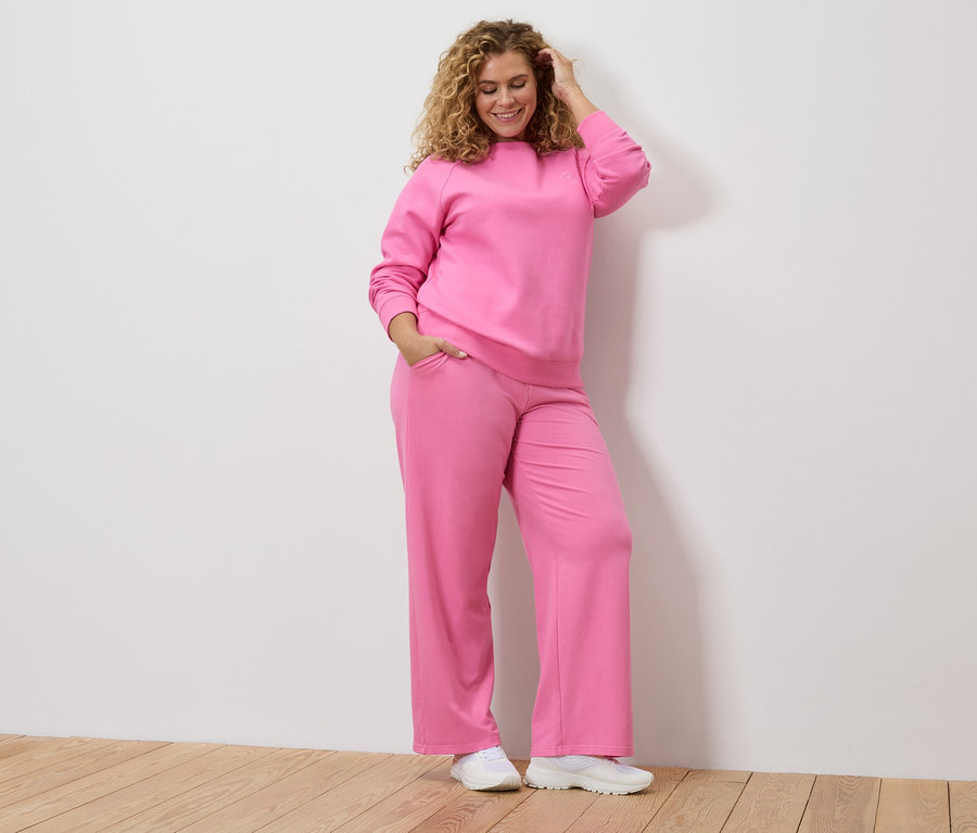 Göndör hajú nő rózsaszín tréningruhában és fehér tornacipőben, kezét a fején tartja.