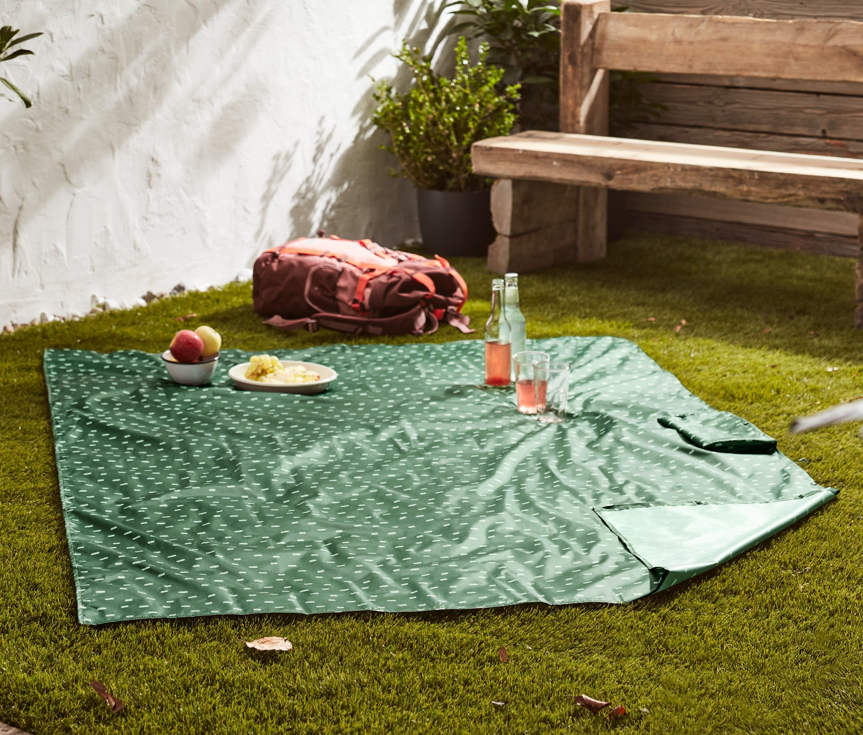 Egy zöld, fehér mintás 2 az 1-ben pikniktakaró terül el a füvön. A takarón tányérok étellel és italokkal.