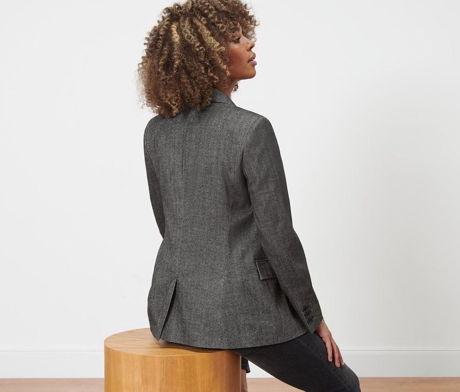 Nő ül egy fa zsámolyon és szőtt blézert visel halszálkás hatással.