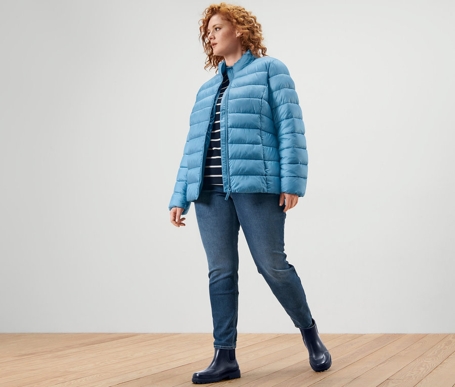 Nő visel steppelt világoskék dzsekit, 3/4-es ujjú felsőt és Fit Nelly treggingst.