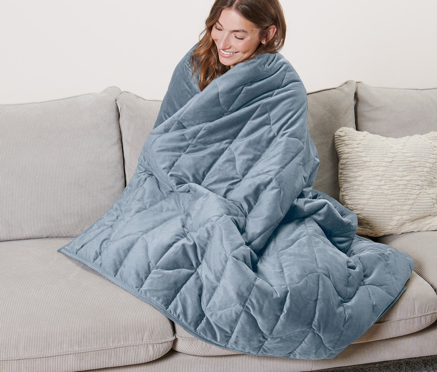 Nő ül a kanapén, kék, 8 kg-os lazító hatású súlyozott takaróba burkolózva.