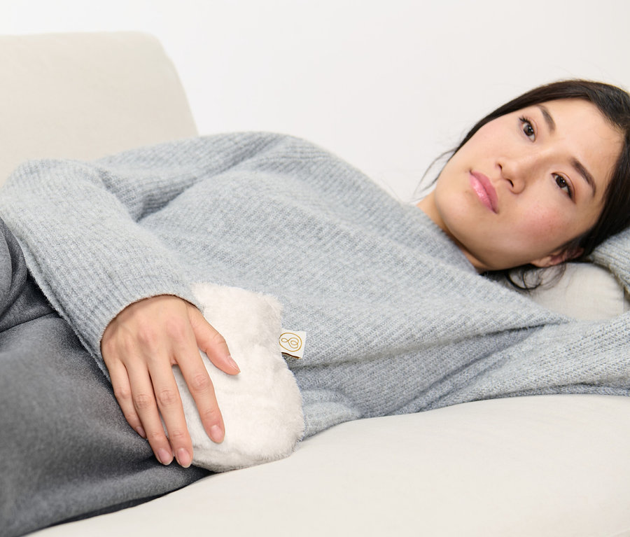 Nő fekszik a kanapén, és egy mini melegvizes palackot tart.