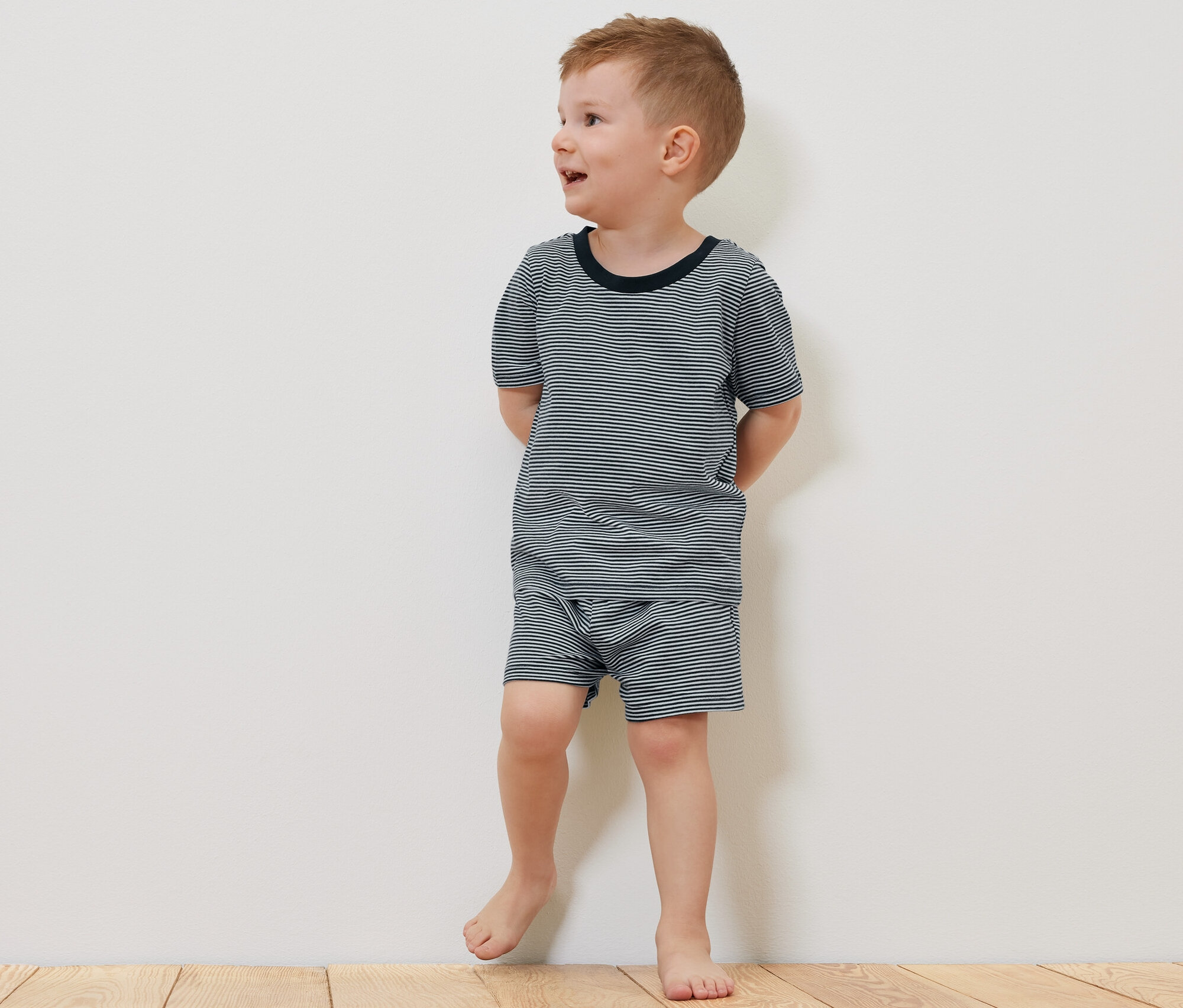 Egy fiú rövid ujjú 2 gyerekpizsama, rövid pizsamában mezítláb áll egy fapadlón egy plüssmedve mellett.