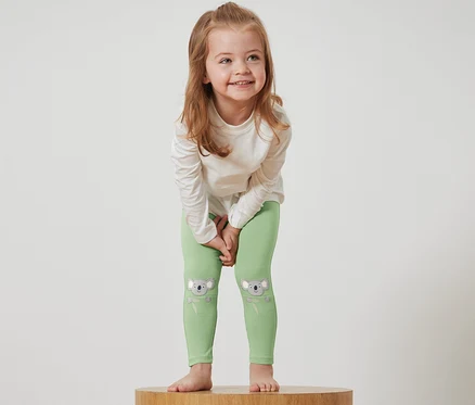 Egy lány áll egy fa talapzaton, zöld, koalás leggingset és fehér pólót visel.