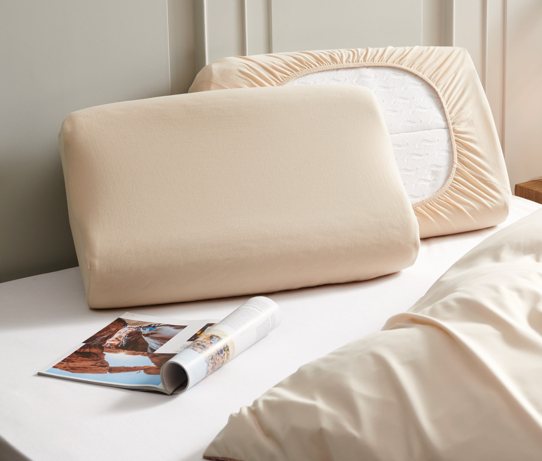 Két bézs színű dzsörzé gumis párnahuzat fekszik egy ágyon egy magazin mellett.